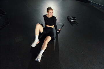 A woman relaxes on a bench in a gym, a contented smile playing on her lips, with dumbbells in the background.