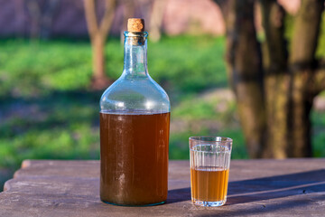 Big bottle with a drink made from fermented birch sap on the wooden table on a warm spring day, closeup. Traditional Ukrainian cold barley drink kvass in a glass jar and glass in yard - 788711806