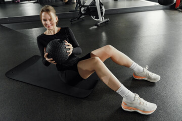 A woman with a wry smile performs a seated exercise on a gym mat, a medicine ball firmly grasped,...