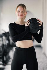With a confident smile, a woman holds a medicine ball at chest height, exuding fitness and poise in her gym attire.