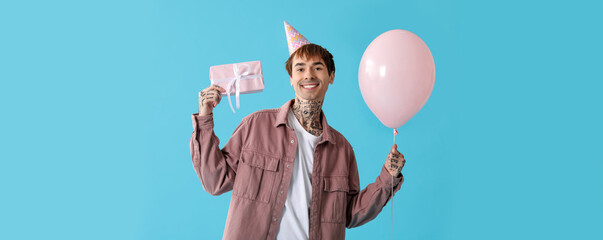 Young man in party hat with gift box and air balloon celebrating Birthday on blue background