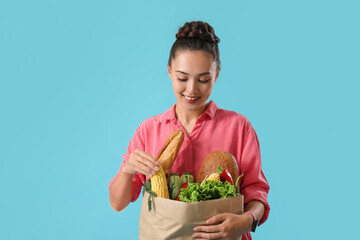 Beautiful young Asian woman holding paper bag with fresh products on blue background