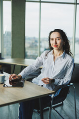 A woman in a white shirt sits at a wooden table and works on a tablet. A young woman works in a modern office