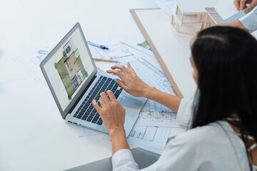 Beautiful caucasian architect using laptop while smart coworker measures house model with...