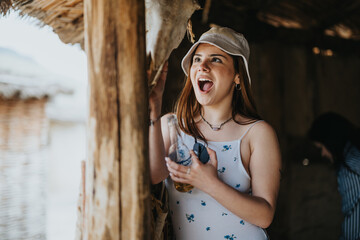 A cheerful young woman wearing a bucket hat and summer dress laughs joyously, holding a glass while sheltered in a rustic beach hut.