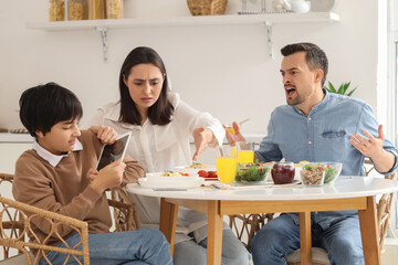 Teenage boy using tablet computer and his angry parents at table during dinner in kitchen. Family problem concept
