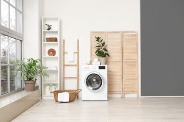 Modern washing machine with basket, shelving unit and dressing screen near white wall. Interior of...