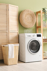 Modern washing machine with basket, shelving unit and dressing screen near green wall. Interior of home laundry room