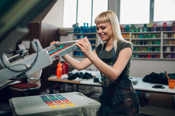 Smiling print shop worker silkscreen printing on t-shirt at facility.