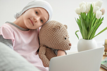 Little girl after chemotherapy with toy bear using laptop at home, closeup. International Childhood...