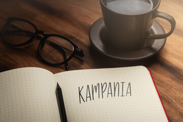 A handwritten inscription "Kampania" on a grille of an open notebook on a wooden countertop, next to a black pencil, a cup with coffee and glasses, a flash of light. (selective focus)