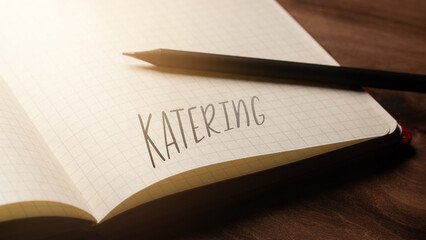 A handwritten inscription "Katering" on a grille of an open notebook on a wooden countertop, next to a black pencil, lighting of light. (selective focus), translation: catering