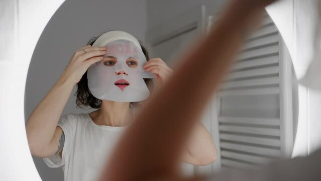 Young woman applies a biocellulose mask to her face at home in her bathroom. Caring for and recovery the skin after cosmetic procedures. Girl relaxation after a difficult day. 