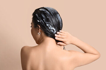 Young woman applying hair mask on beige background, back view