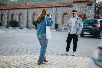 A young woman in denim captures her stylish friend with sunglasses outdoors near a historic building, depicting a casual, joyful day.