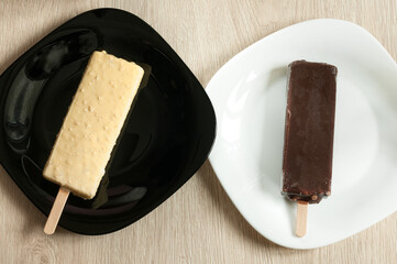 Two ice creams on sticks in dark and white chocolate glaze on different plates. View from above