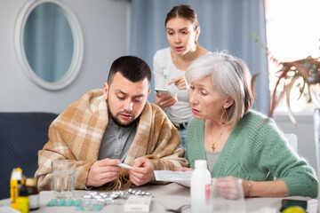 Sick man having viral disease, young and elderly anxious female relatives choosing medicines to help him