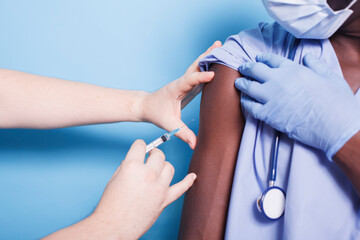 African american nurse wearing face mask, getting vaccinated with syringe and needle. Close-up of medical assistant receiving vaccine shot from hands of doctor for virus protection. - obrazy, fototapety, plakaty