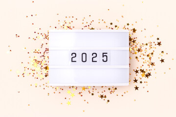 Lightbox with 2025 numbers with frame made of golden stars confetti on a beige background. New Year creative concept.