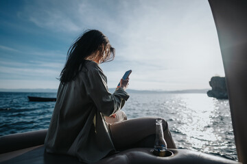 A happy young girl relaxes on a boat, holding a smart phone with a joyful expression, surrounded by the vast sea under a clear sky.