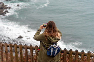 Woman with a backpack on her back taking a picture with her mobile phone of the landscape of the touristy Basque coast on a cloudy day.