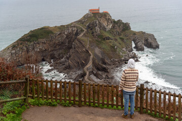 Man with his back turned watching the hermitage of San Juan de Gaztelugatxe located on a hill in the middle of the sea on the touristy coast of Biscay.