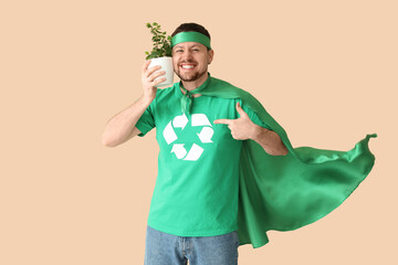 Young man dressed as eco superhero with houseplant pointing at recycling logo on his t-shirt against beige background. Ecology concept