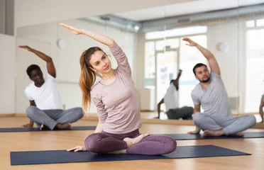 Foto op Canvas Positive sporty young woman doing stretching exercises while sitting in lotus position during training with group of people in fitness center © JackF