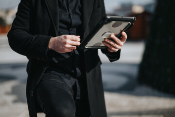A modern, well-dressed male professional engaging with a digital tablet for business tasks outside in a sunny urban setting.