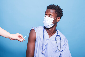 Image of medical nurse getting vaccinated by doctor in studio. Medic using syringe with needle for vaccine shot on arm of healthcare assistant. Specialist receiving vaccination against coronavirus. - obrazy, fototapety, plakaty
