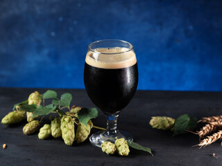 A glass of beer stout or porter on a dark blue background, green cones of hops