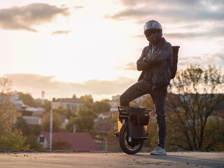 A young male rider in protective gear with a backpack on an electric unicycle against the background of the city at sunset. Driving around the city on a mono wheel