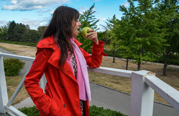 Brunette woman wearing a red coat  eating a green apple, standing beside a white railing with trees and a park in the background. Scene juxtaposing vibrant and natural colors in an everyday moment