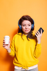 Portrait of female adult grasping mobile device and cup of coffee while using headphones. Caucasian woman enjoying music on wireless headset while using smartphone and having drink.