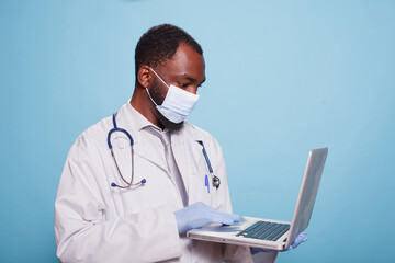 Medical physician with latex gloves and face mask holding a laptop to review findings of a clinical diagnosis. Portable computer is being held by medic wearing lab coat and stethoscope.