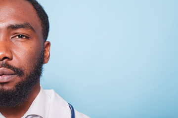 Close-up of African American doctor with a beard looking at the camera. Half-face portrait of black male healthcare specialist wearing white lab coat against isolated blue background.