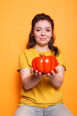 Fototapeta na wymiar Portrait of caucasian woman gripping a bell pepper and looking into camera standing in studio with orange background. Young lady having healthy diet, eating vegetables. Organic veggie concept.