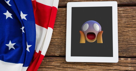Fototapeta premium A tablet displaying shocked emoji rests on a wooden surface beside American flag
