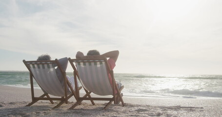 Naklejka premium Senior biracial couple relaxing on beach chairs by sea