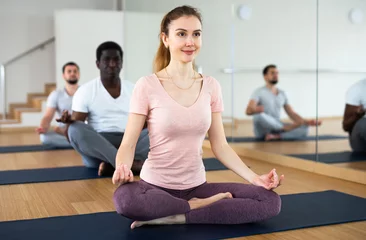 Tuinposter Young positive woman sitting in lotus pose on mat in fitness room, doing yoga meditation with group of adult people.. © JackF