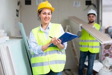 Smiling asian female civil engineer in yellow safety vest and hard hat standing with documents in...