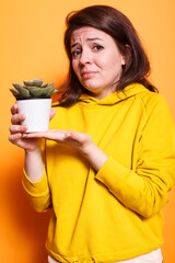 Female plant lover displaying green plant in a white pot on camera, holding natural decoration in hands. Caucasian woman having a tropical vegetation and greenery in palms.