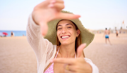 Portrait cheerful of attractive Caucasian woman with hat posing for photo doing hand frame gesture on beach. Smiling female looking through fingers at camera. Gen z joyful people summer travel outdoor