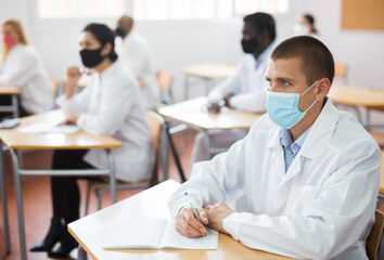 Focused adult medical student in protective face mask studying in classroom with group, writing...