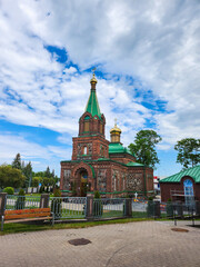 A charming small, old Russian Orthodox Church with a green roof and golden domes nestled among tall green trees under a clear blue sky with fluffy white clouds creating a serene and picturesque scene.