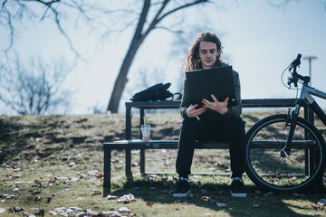 Handsome young man with long hair sits on a park bench, working on a laptop, with his bicycle beside him on a sunny day.