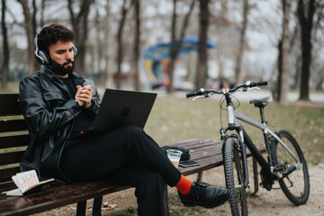 Stylish male entrepreneur remotely working with headphones and laptop in a city park next to his bicycle.