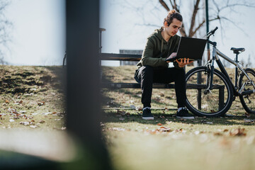 A good looking male enjoys a relaxing weekend outdoors, working on his laptop while sitting next to his bicycle in a sunny park.