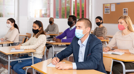 Group of students in protective mask listening attentively to teacher explaining material in classroom