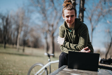 A young adult male enjoys a sunny day in the park, working on his laptop with his bicycle resting beside the bench.